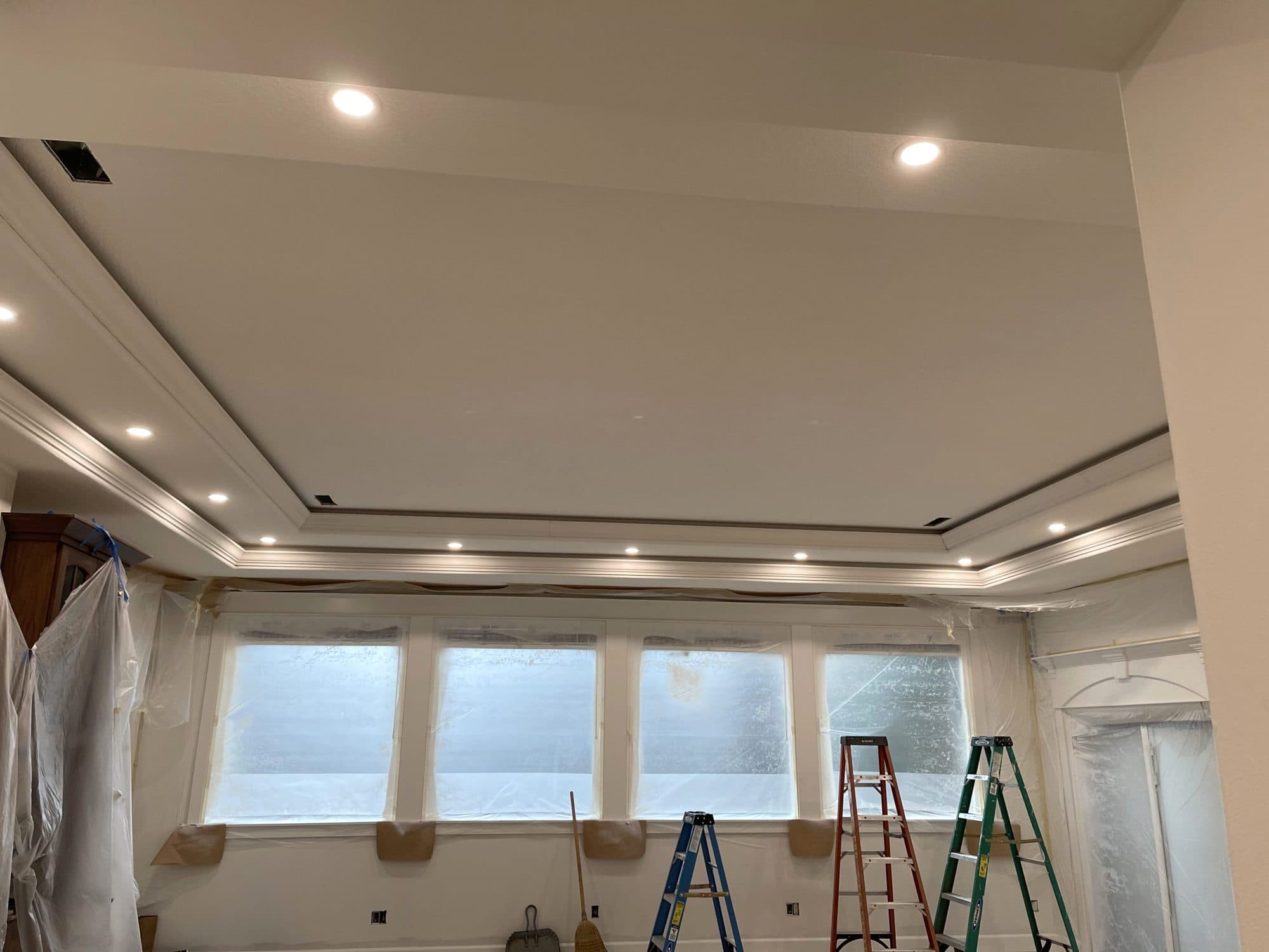 A living room ceiling mid-job, with new recessed lights set around a tray ceiling and the windows still covered in plastic.