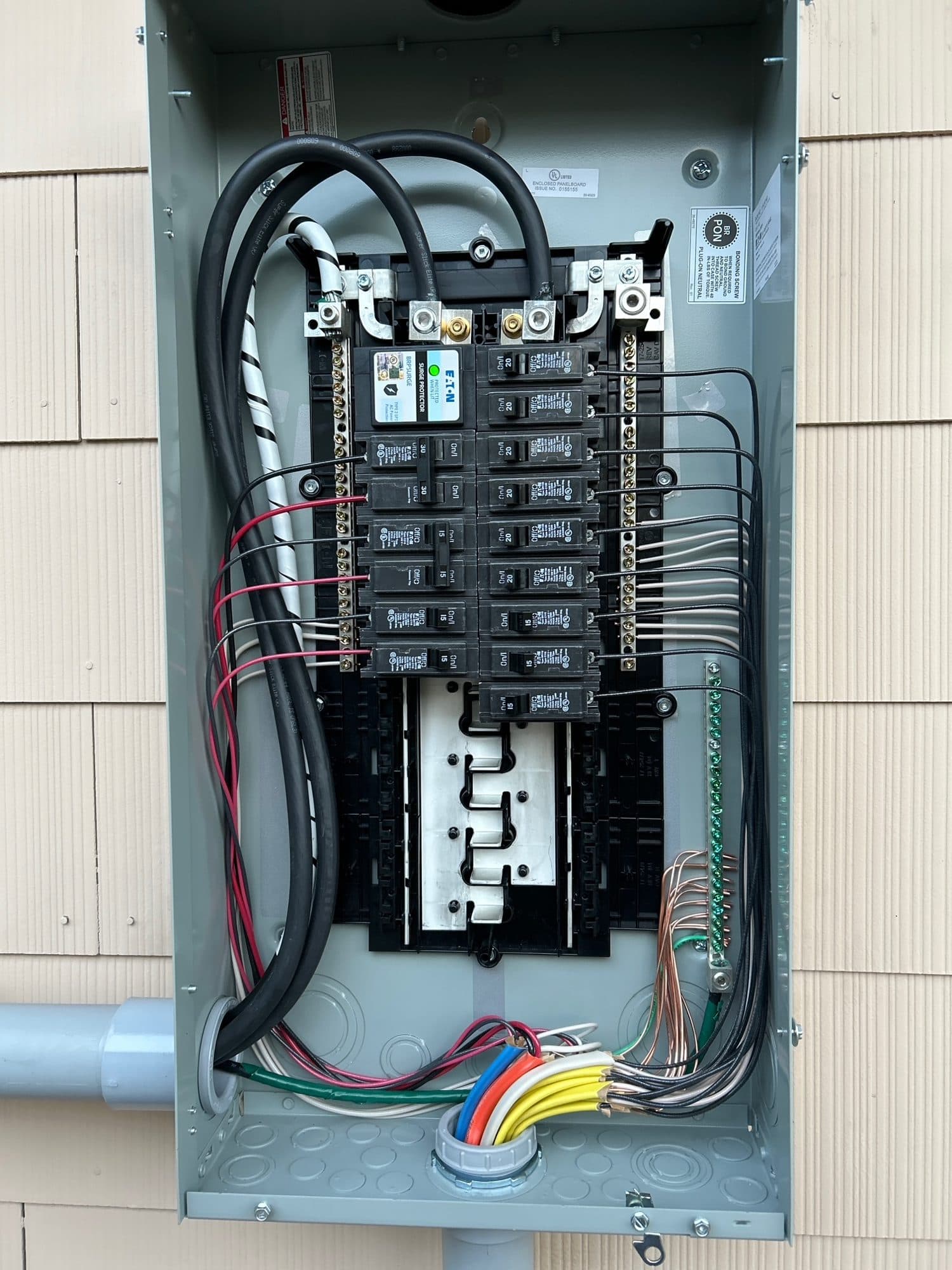 Rows of new breakers in a gray outdoor panel on a shingle-sided house, with the new wires entering through the bottom.