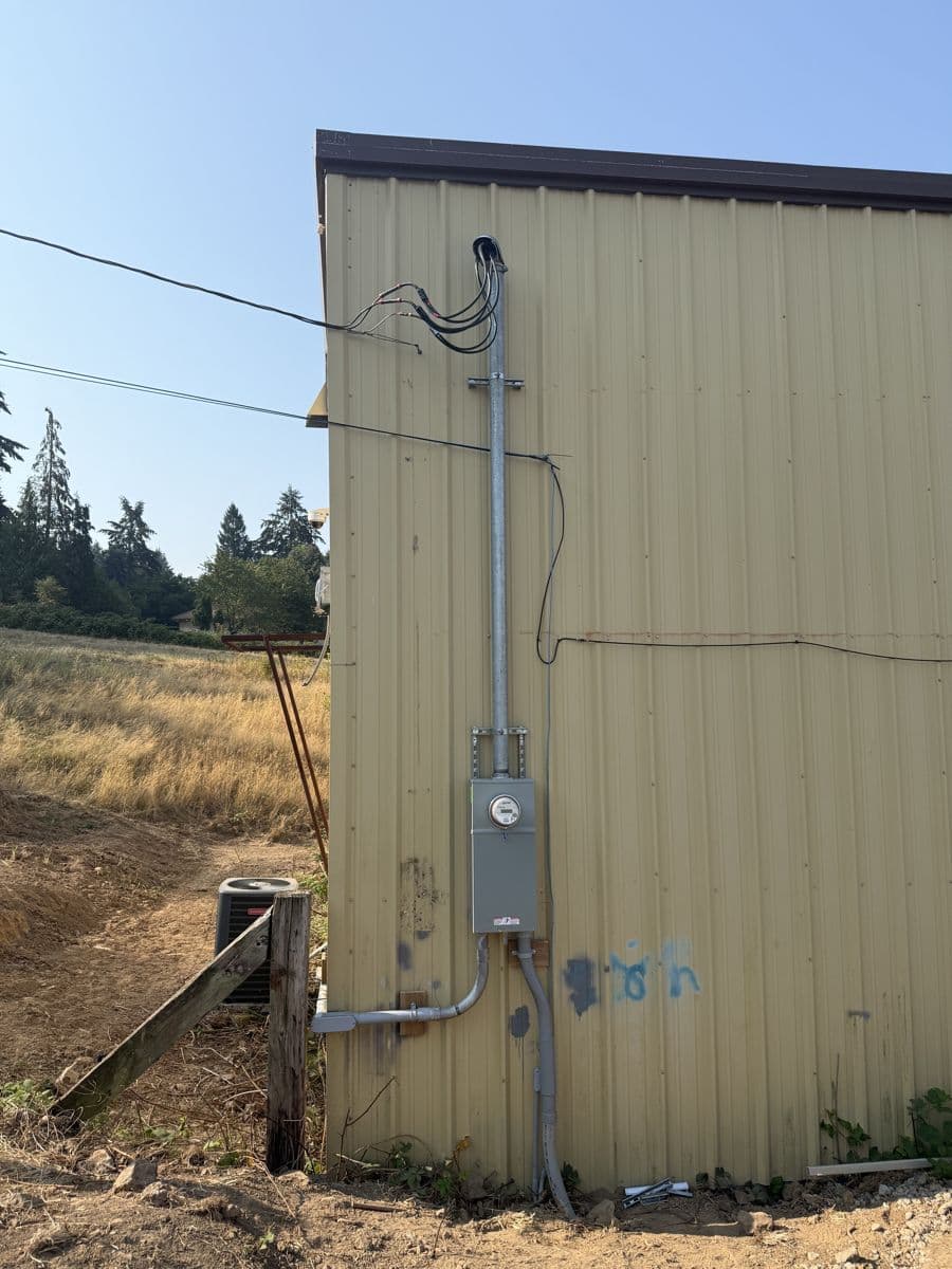 A new service mast, meter base and gray conduit on the side of a metal outbuilding, with the overhead wires connected at the top of the mast.